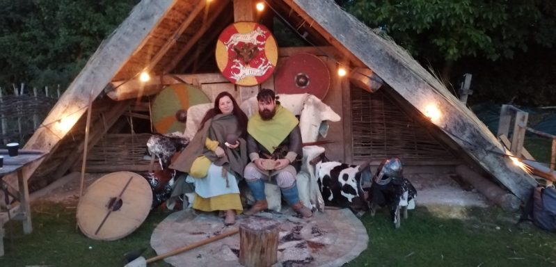 Two reenactors dressed in Viking clothing sit surrounded by shields and furs.