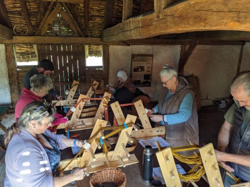 A group of course participants stand weaving at inkle looms in a replica Saxon historic building. 